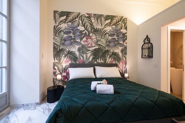 A bed with a green quilt, a floral wallpaper, a black bedside table, and a birdcage on the wall.