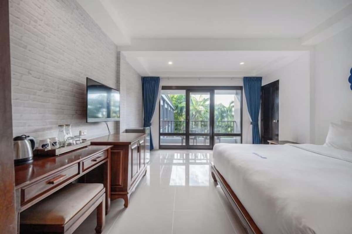 A modern hotel room with a flat-screen TV, a wooden desk with a bench, a large bed, and a balcony with a view of greenery.