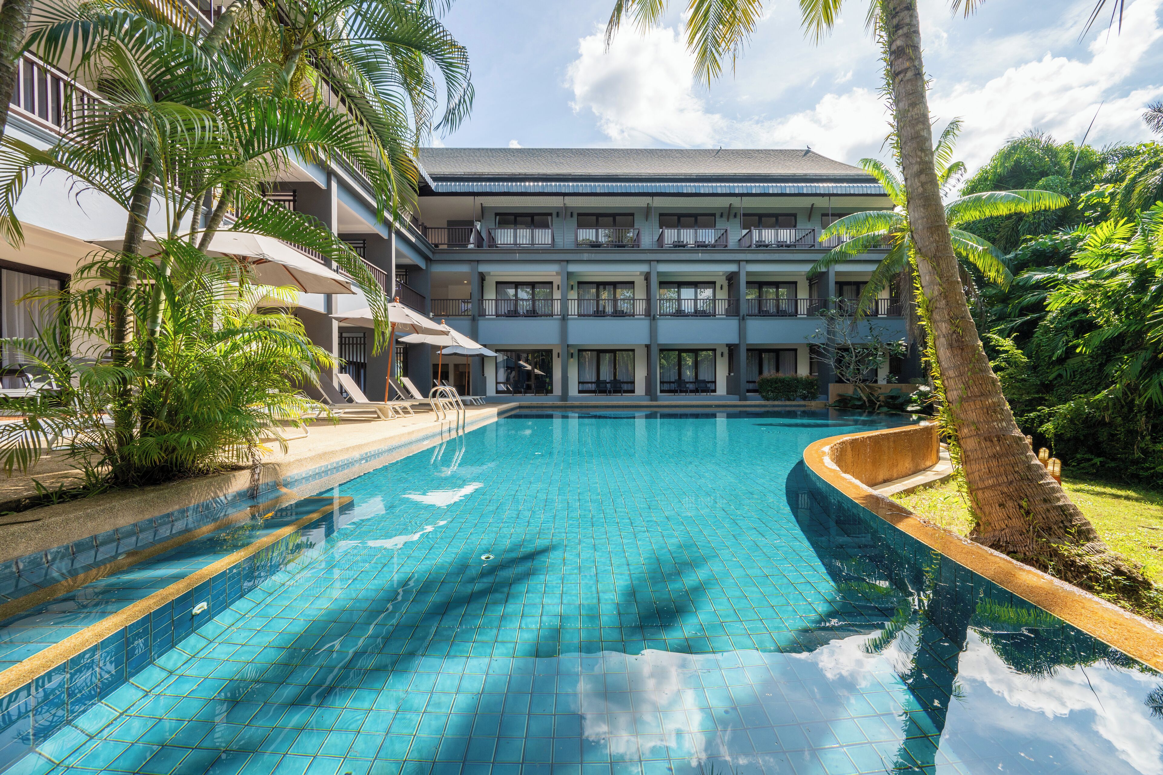 A swimming pool with a tiled edge, surrounded by palm trees and a multi-story building with balconies.