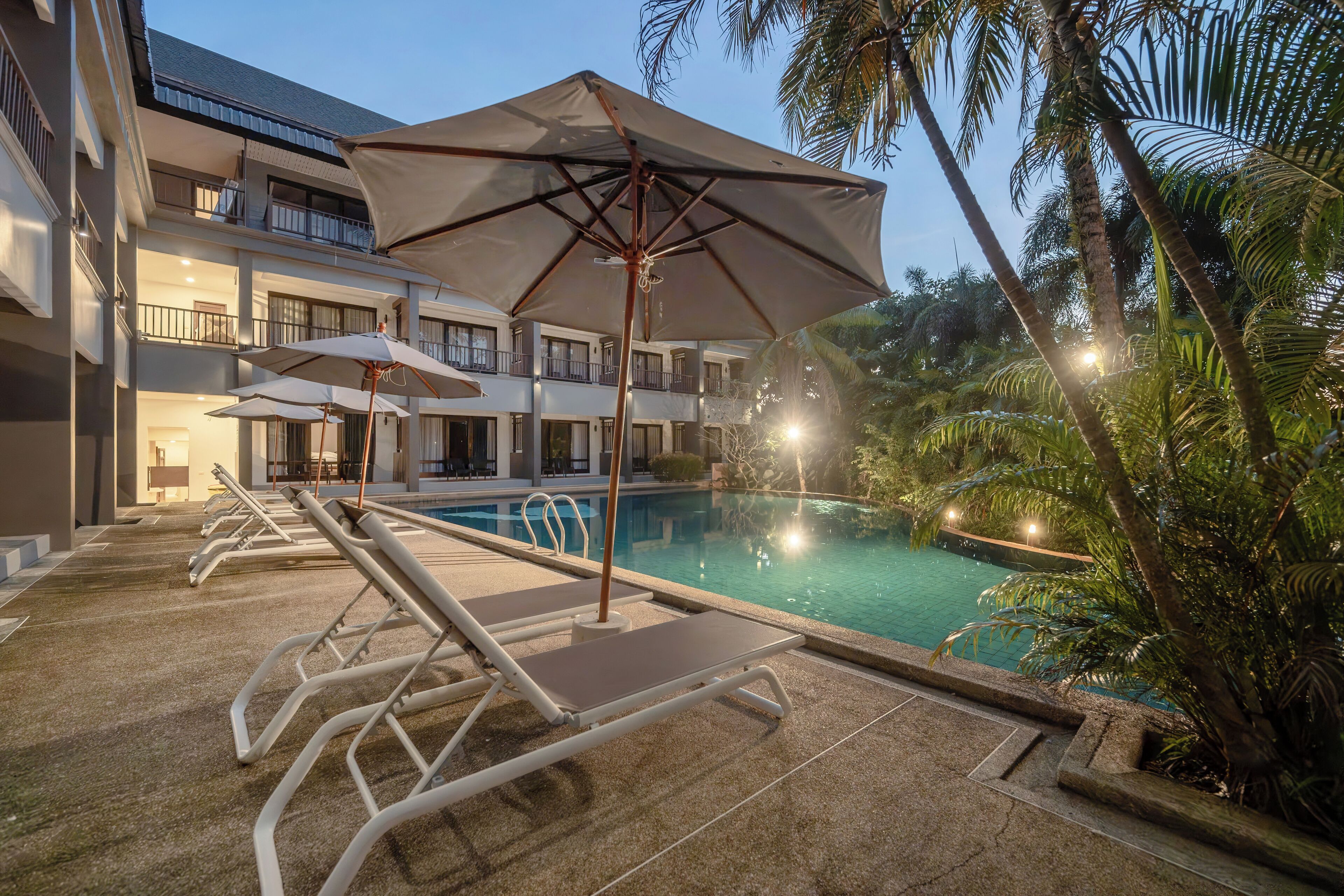 A hotel pool area with lounge chairs and umbrellas, surrounded by palm trees and a building with balconies.