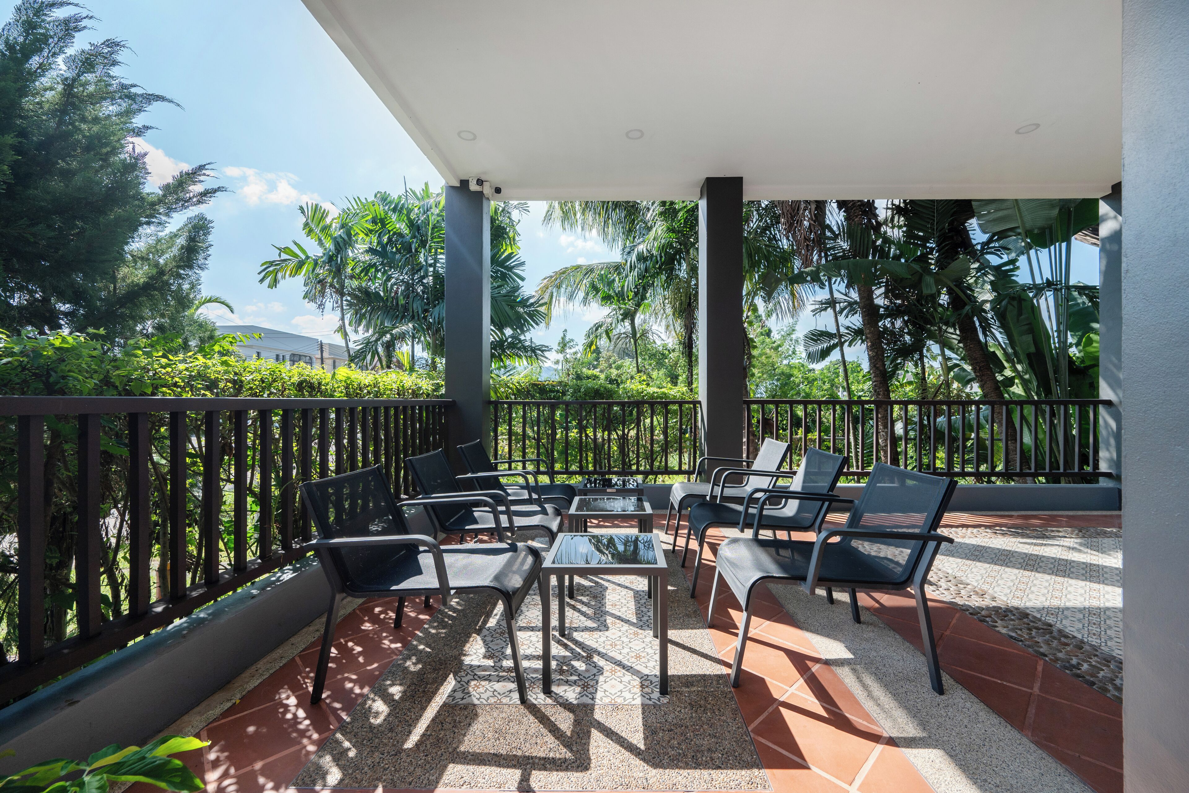 A covered outdoor seating area with chairs and tables, surrounded by a railing, and overlooking a lush garden with palm trees.