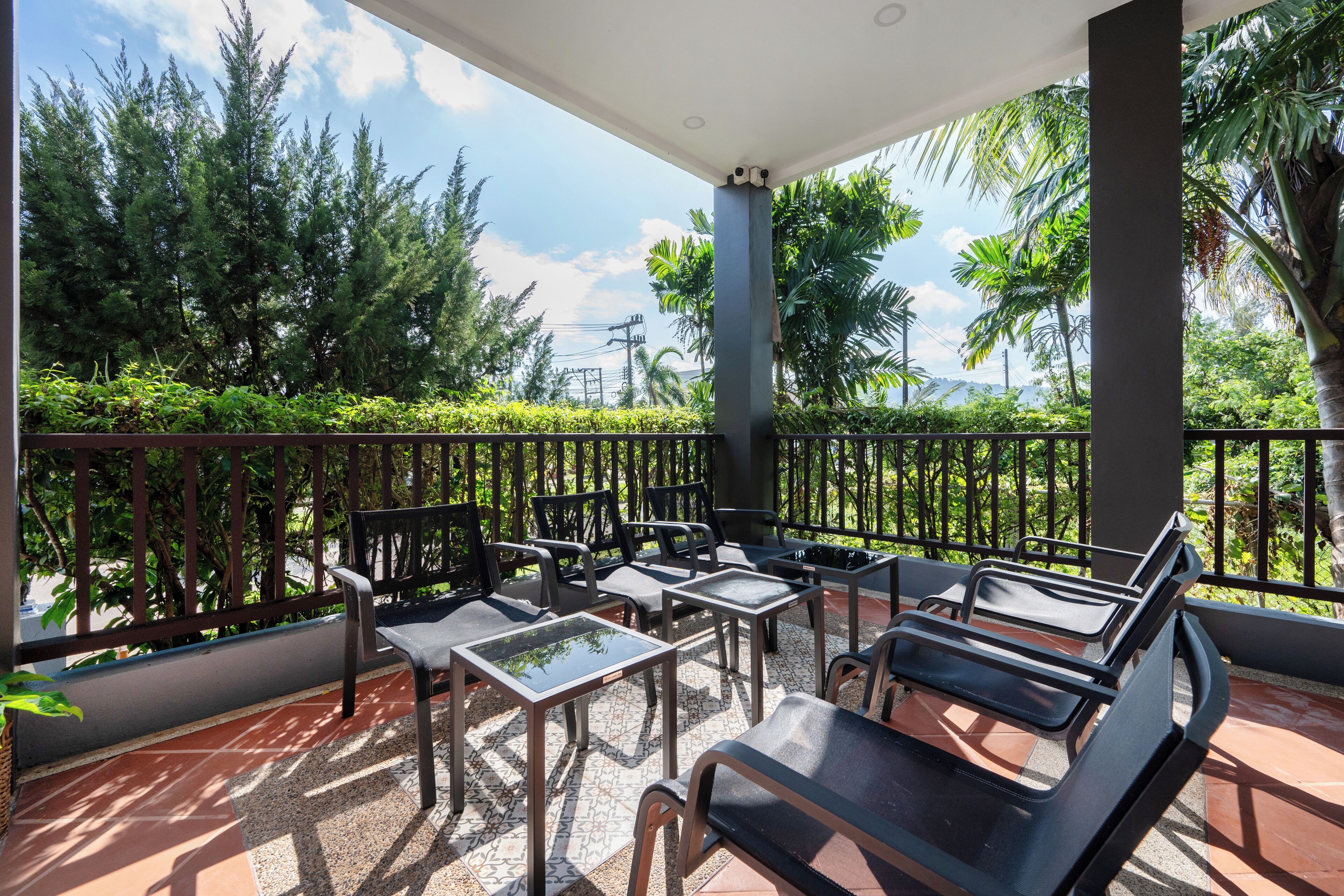 A covered outdoor seating area with tables and chairs, surrounded by a metal railing, and overlooking a lush garden with various trees and plants.
