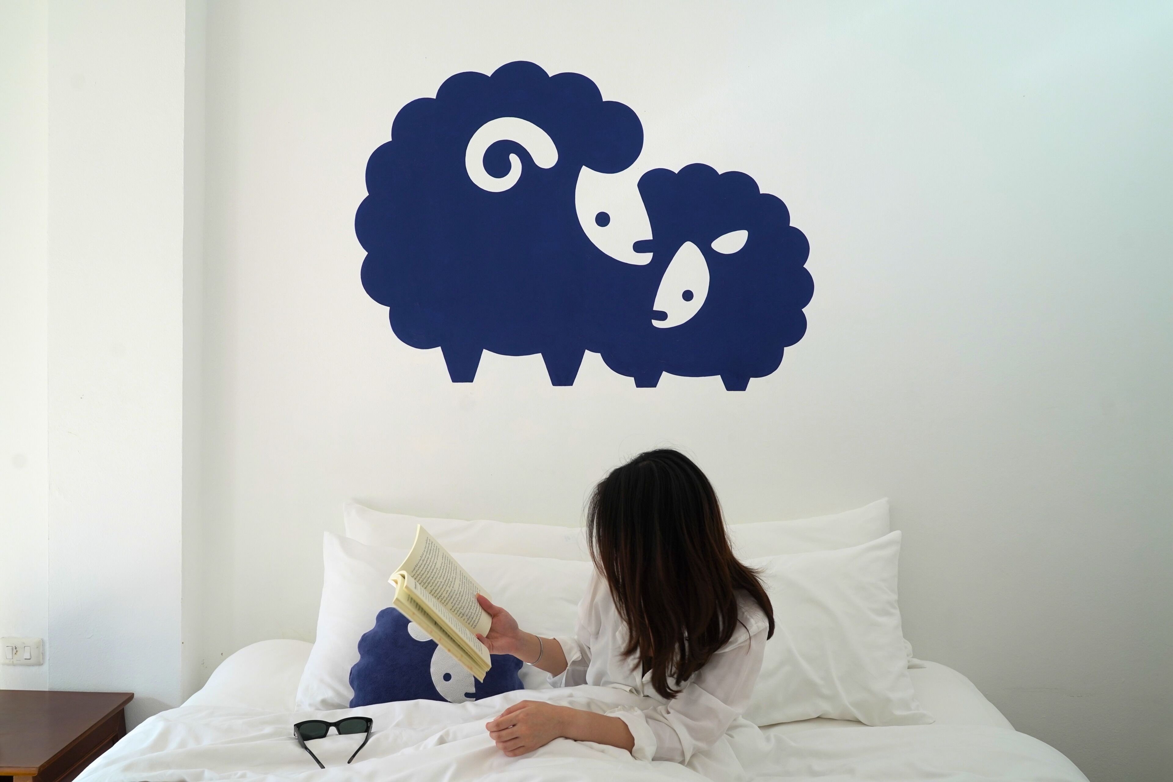 Une personne est assise dans une pièce et lit un livre. Un mur de la pièce est décoré d’un motif représentant des moutons.