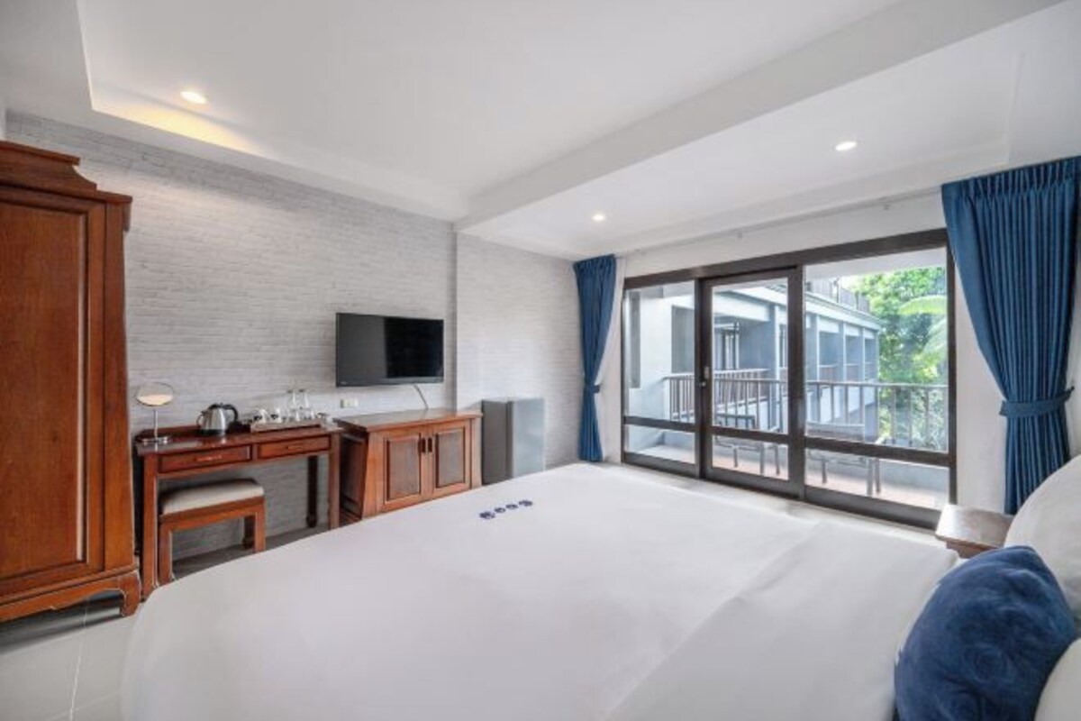 A hotel room with a large bed, a desk with a chair, a flat-screen TV mounted on the wall, and a balcony with a view of the outside.