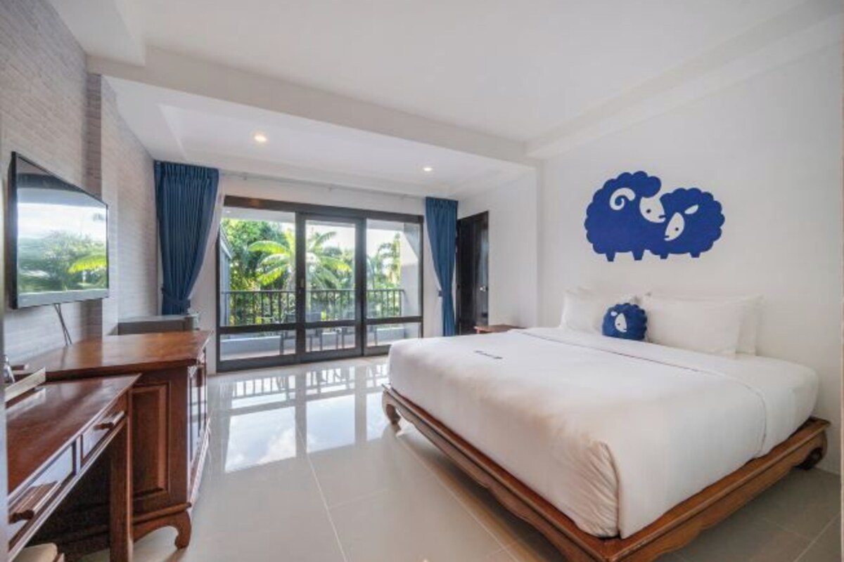 A hotel room with a large bed, a TV, a wooden dresser, and a balcony with a view of greenery.