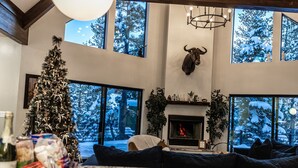A cozy living room with a Christmas tree, a fireplace, and large windows overlooking a snowy landscape. - Duck Creek Luxury Cabin w/ Hot Tub | Las Hermanas (Kane County)
