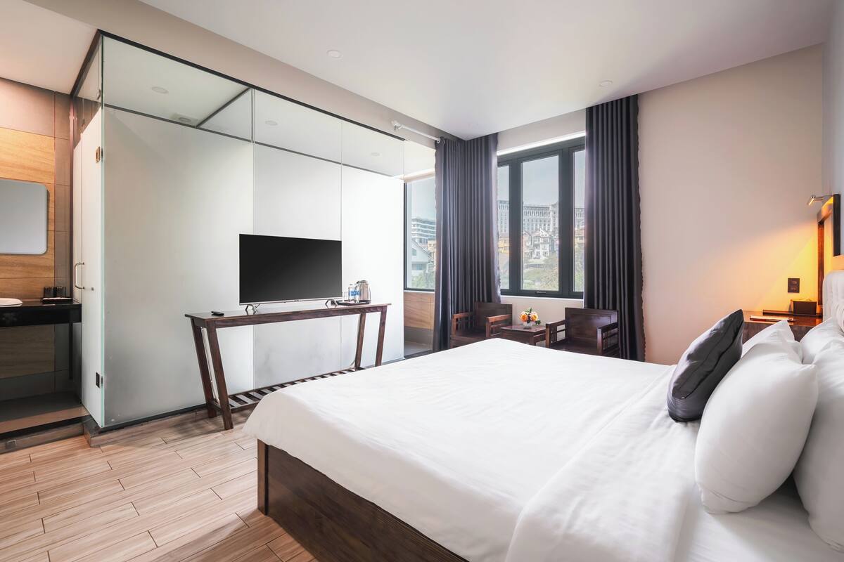 Habitación de hotel con una cama grande, un escritorio con televisor de pantalla plana, una silla y vistas a la ciudad a través de amplias ventanas.