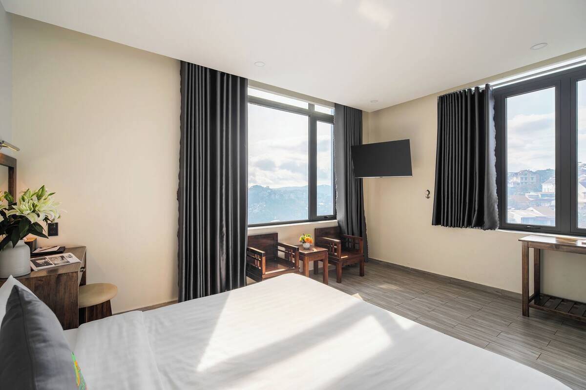 Habitación de hotel con una cama grande, un escritorio con una silla, una mesita con una planta, un televisor montado en la pared y una ventana con cortinas.