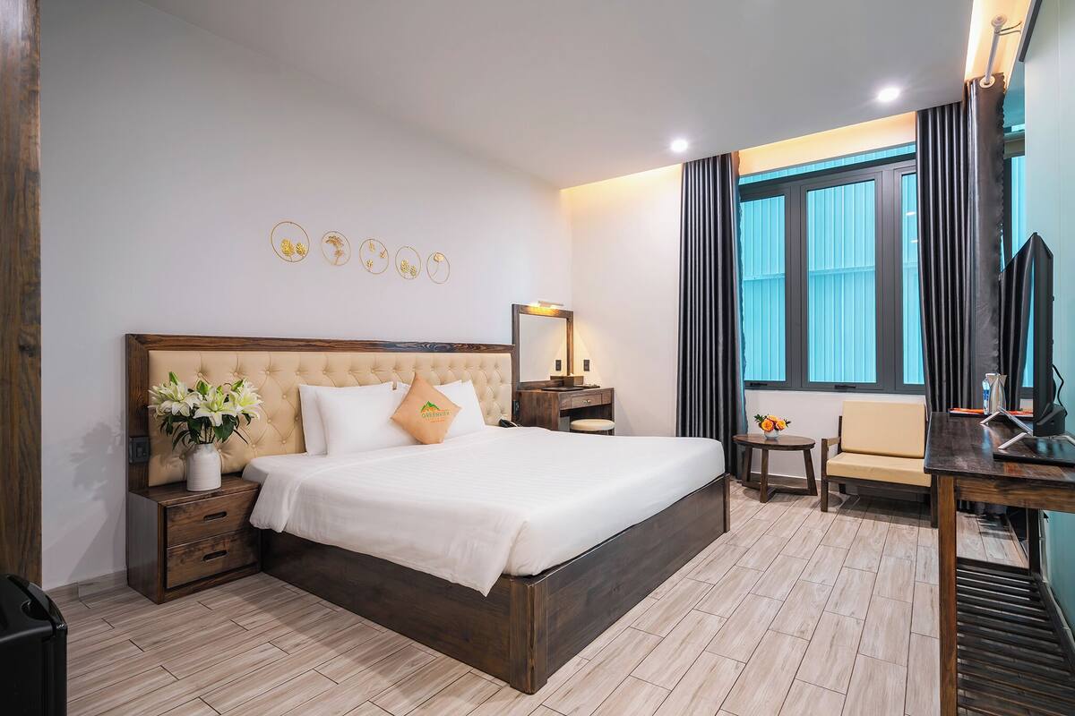 Una habitación de hotel con una cama grande, una mesita de noche con lámpara, una pequeña mesa con un florero y una ventana con cortinas.