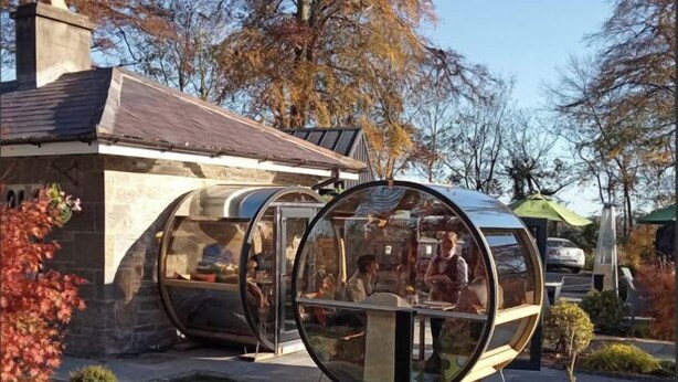 Een glazen constructie met een tafel en stoelen, gelegen in een tuin met bomen en een gebouw op de achtergrond.