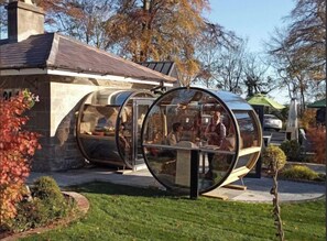 A glass-enclosed structure with a table and chairs inside, situated in a garden with trees and a building in the background.