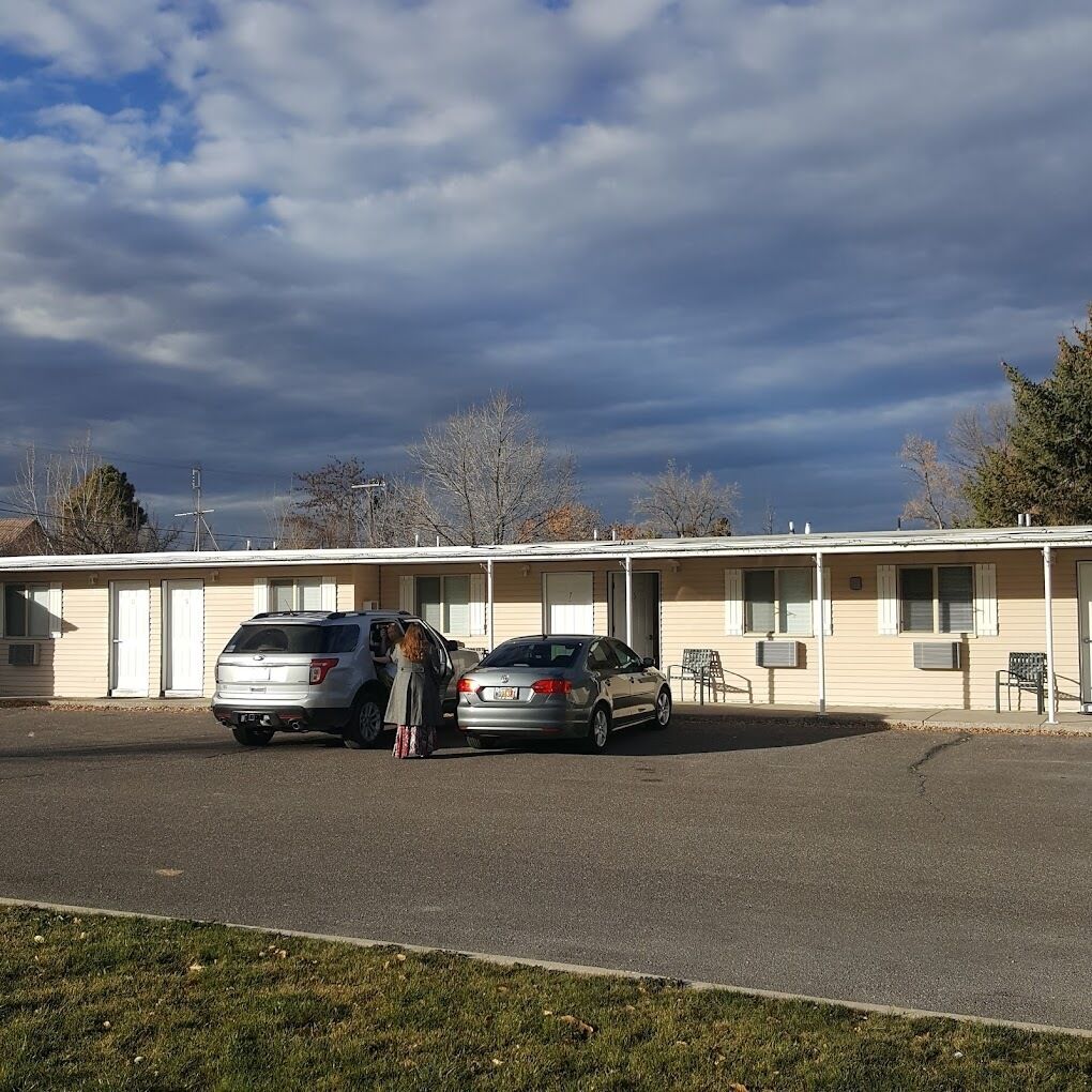 Un motel avec deux voitures garées devant, une pelouse au premier plan et un ciel dégagé avec quelques nuages.