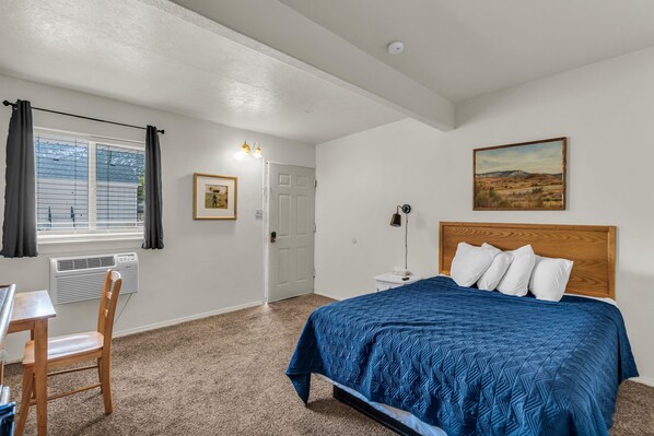 A bedroom with a bed, a desk, a chair, a window with curtains, a door, and a painting on the wall.