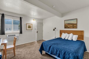 Un dormitorio con cama, escritorio, silla, ventana con cortinas, puerta y un cuadro en la pared.