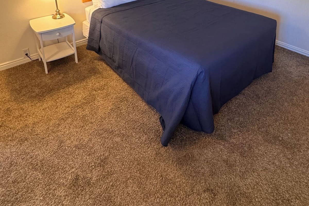 A neatly made bed with a dark blue bedspread and white pillows, a wooden headboard, a small bedside table with a lamp, and a carpeted floor.
