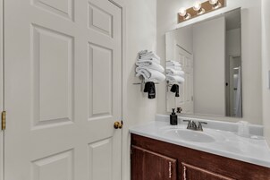 Un baño con una puerta blanca, un lavamanos con un espejo encima y toallas colgadas en un perchero.