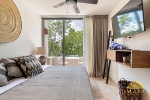 Ein Schlafzimmer mit Bett, Fernseher, Fenster mit Vorhängen, Balkon mit Tisch und Stühlen sowie einem Wandregal.
