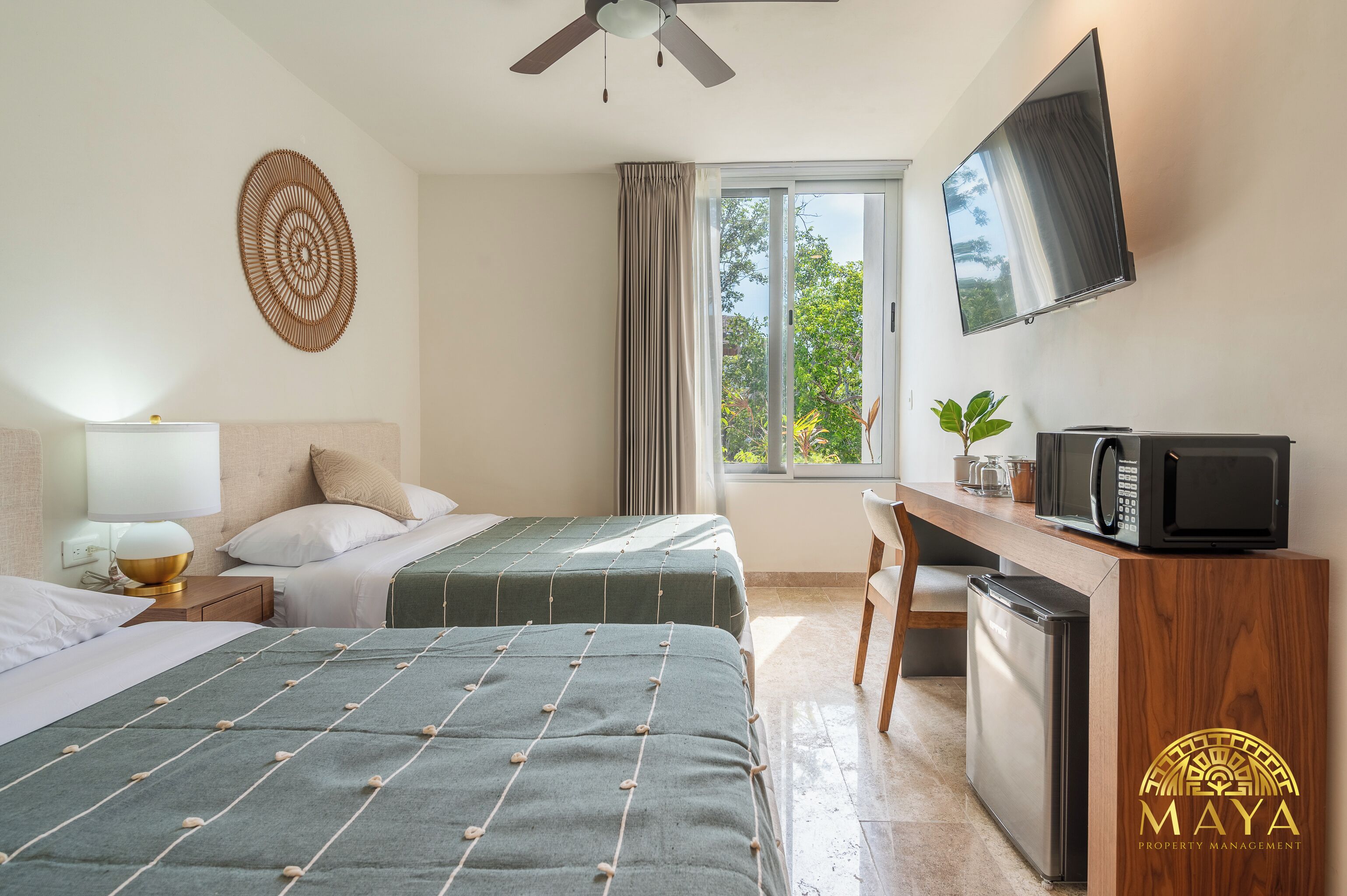 A hotel room with two beds, a desk with a TV, a microwave, and a view of trees through the window.