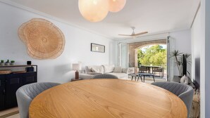 Ein modernes Wohnzimmer mit einem runden Holztisch, grauen Polsterstühlen, einem Deckenventilator und Blick ins Freie durch eine Glastür.