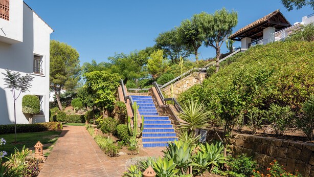 Ein Weg führt an einer steinernen Treppe entlang, umgeben von üppiger Vegetation und einem weißen Gebäude mit einem Ziegeldach.