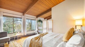 A bedroom with a large bed, a chair, a small table, and a view of trees through large windows. - Perch - Lake Forest Park Retreat (Kenmore)