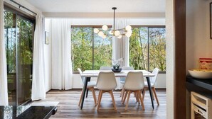 A modern dining area with a white table and chairs, a chandelier, and large windows with a view of trees. - Perch - Lake Forest Park Retreat (Kenmore)