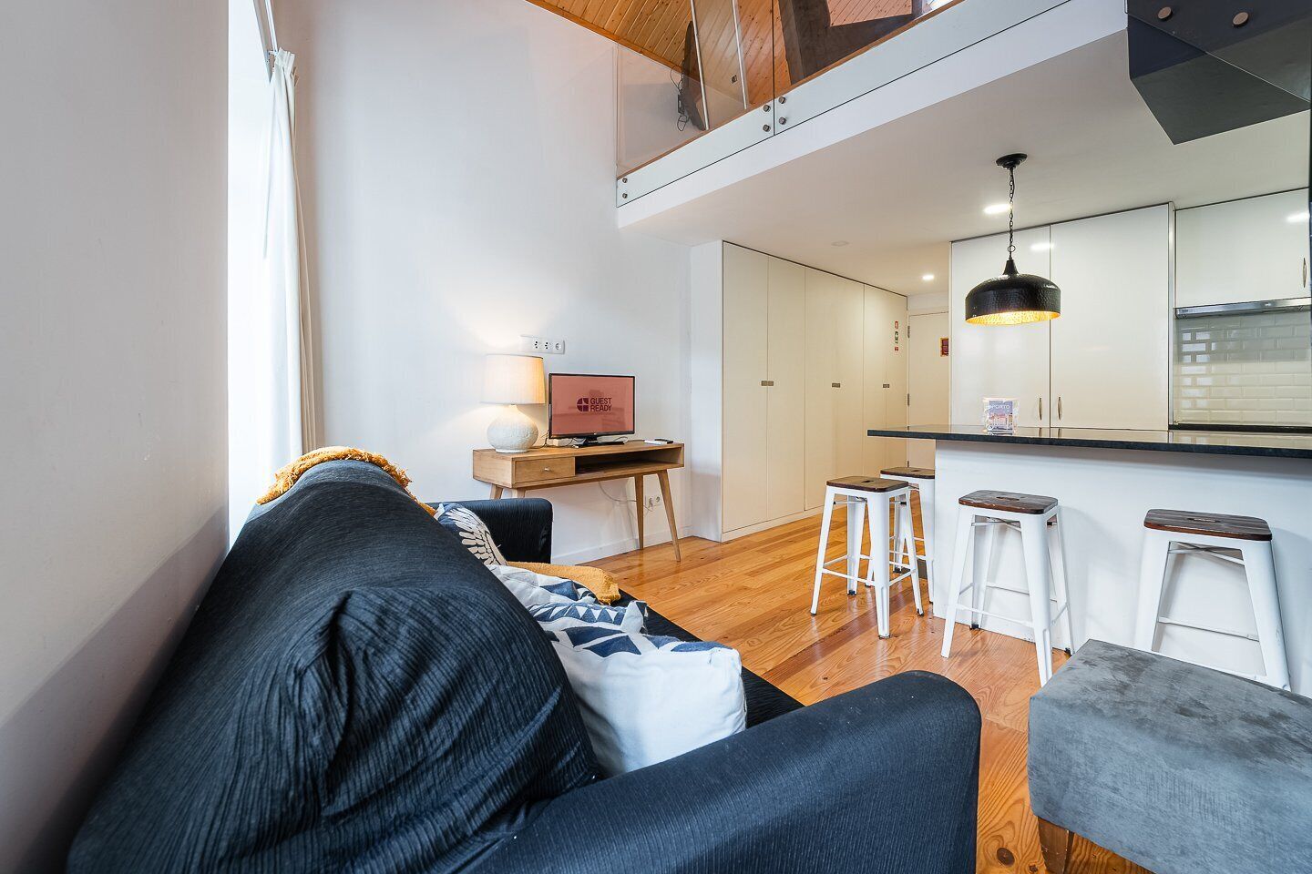 A modern living room with a sofa, a desk with a computer, and a kitchen area with stools.