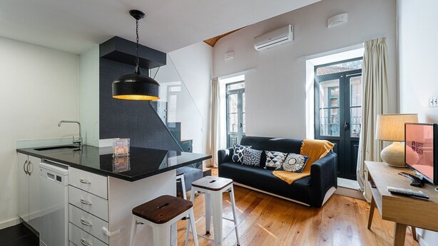 A modern kitchen with a black countertop, white cabinets, and a hanging pendant light. There is a black sofa with patterned cushions in the living area, a small white stool, and a wooden table with a lamp and a sign that reads "GUEST READY". - GuestReady - Charming mezzanine near Porto (Vila Nova de Gaia)