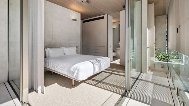 A modern bedroom with a large bed, a ceiling fan, and a view of a balcony with plants. - Hastings Villas (Noosa Heads)