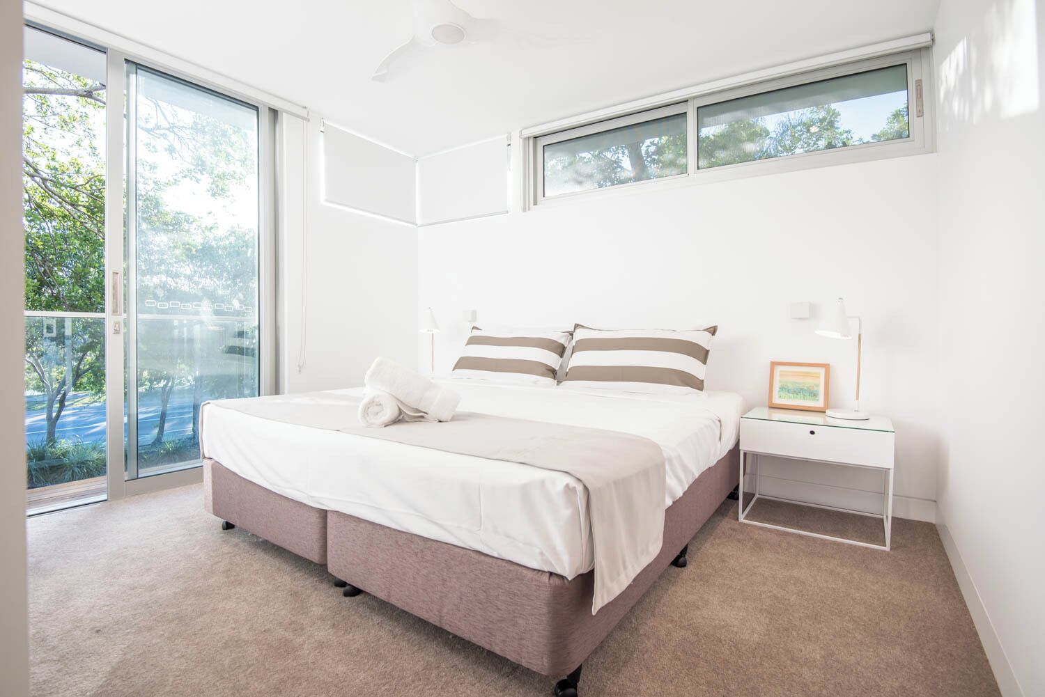 A modern bedroom with a large bed, a bedside table with a lamp, and a view of the outdoors through a sliding glass door.
