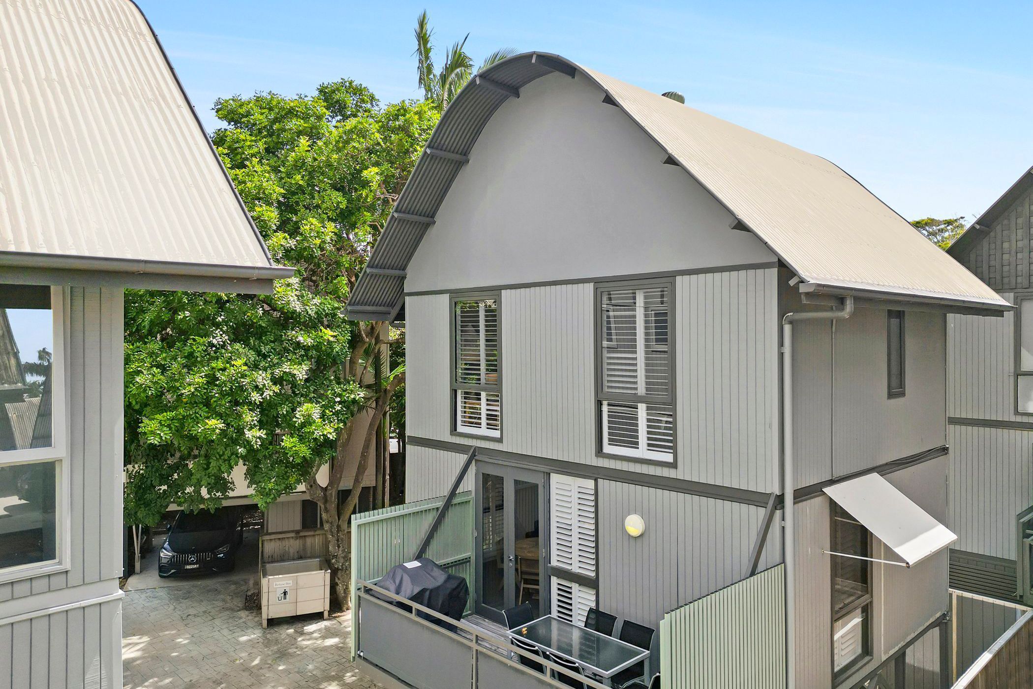 A two-story house with a balcony, a car parked in the driveway, and a tree in the background.