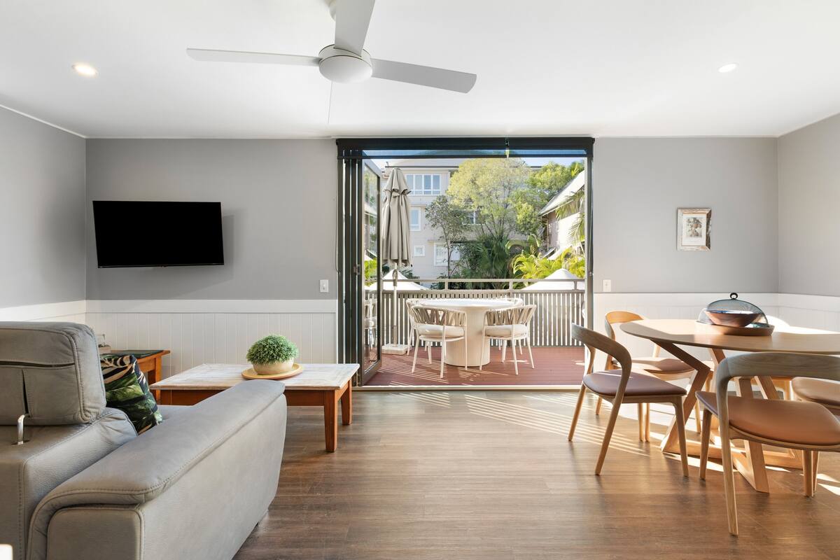 Uma sala de estar moderna com um sofá, uma mesa de jantar com cadeiras, uma TV de tela plana na parede e uma porta de vidro deslizante que dá para um pátio externo.