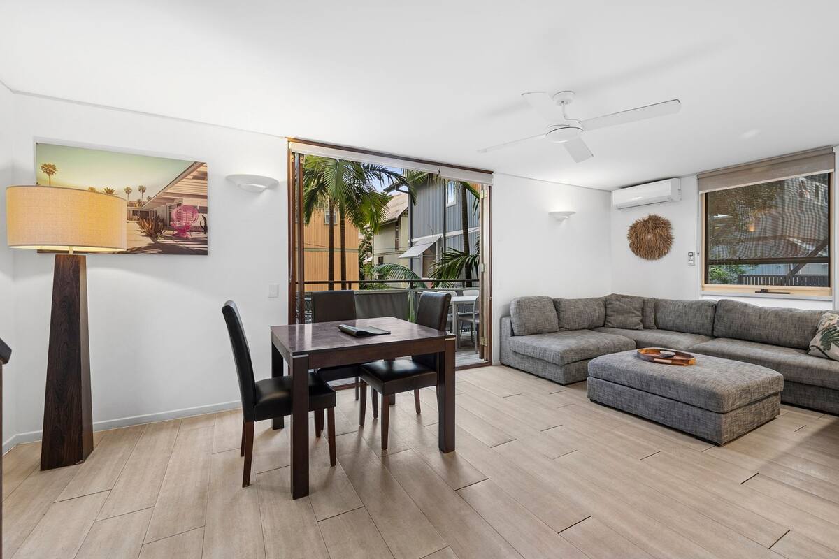 Uma sala de estar moderna com área de jantar, um sofá cinzento, um ventilador de teto e uma janela grande.