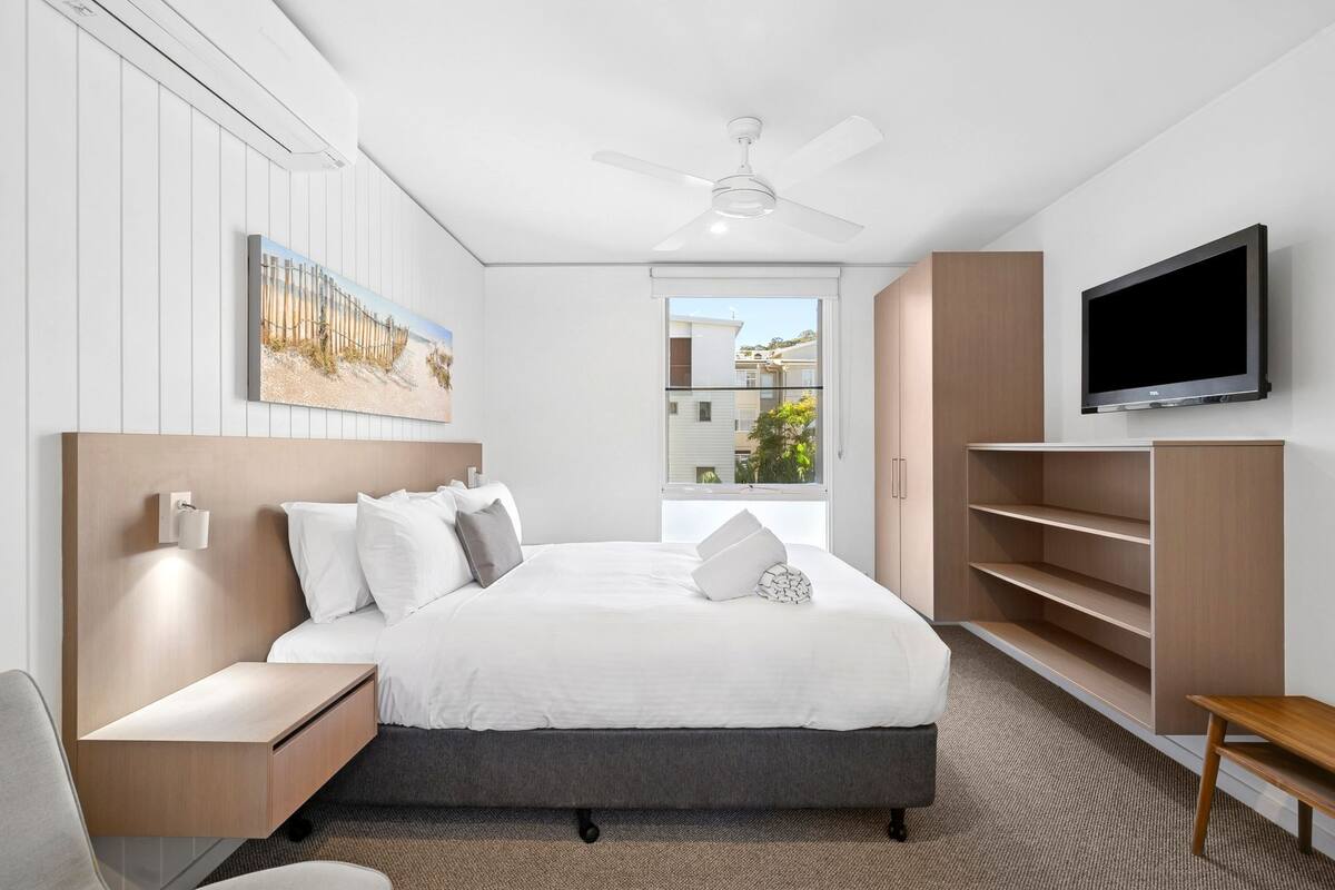 Quarto de hotel moderno com uma cama grande, televisão, ventoinha no teto e vista para o exterior.
