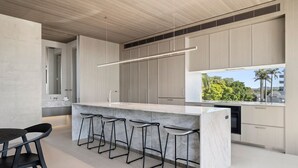 A modern kitchen with a marble island, bar stools, and a large window with a view of greenery. - Hastings Villas (Noosa Heads)