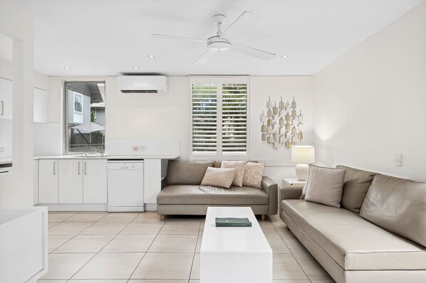 Un salon moderne avec un canapé beige, une table basse blanche et un coin cuisine doté de meubles blancs et d’une fenêtre avec des stores.