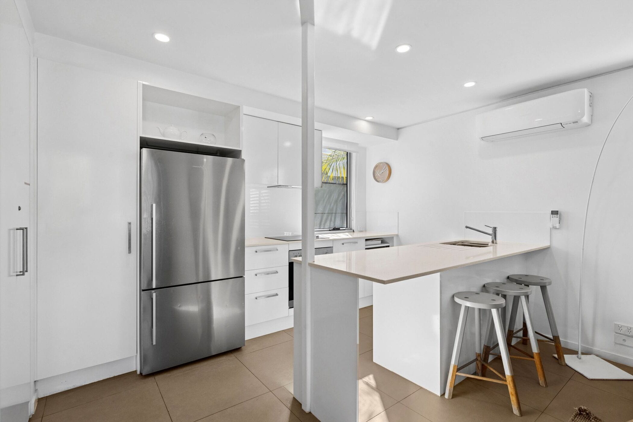 A modern kitchen with white cabinetry, a stainless steel refrigerator, a built-in oven, and a small island with stools.