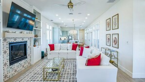 A spacious living room with a fireplace, a large flat-screen TV, a ceiling fan, and a kitchen in the background.