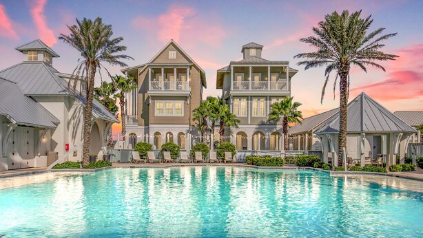A large swimming pool surrounded by two-story buildings with balconies, palm trees, and a sunset sky.