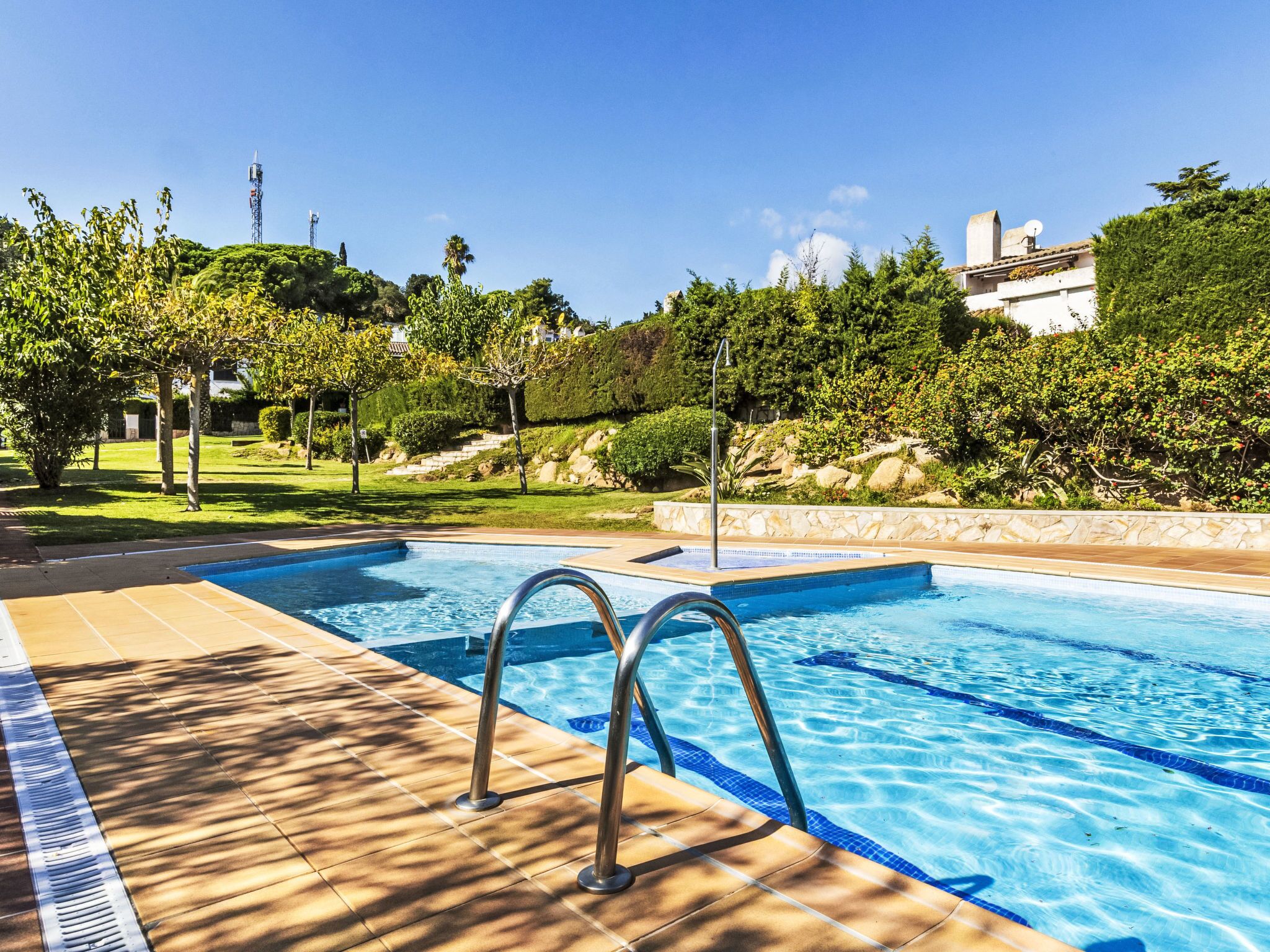 A swimming pool with a tiled deck and metal ladder, surrounded by greenery and a stone wall.