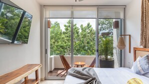 A modern hotel room with a large bed, a wooden bench, a TV, a balcony with a view of trees, and a hanging light fixture. - NHOA 311A Lux 2BR Pool View Aldea Zama (Tulum)