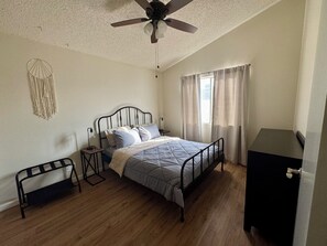 Un dormitorio con cama, ventilador de techo, mesita de noche, una decoración de macramé en la pared y una ventana con cortinas.