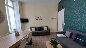 A living room with a dining table, two sofas, a TV, and a wall clock. - Apartment T2+ ( sleeps 3 ) (Bagnères de Luchon)