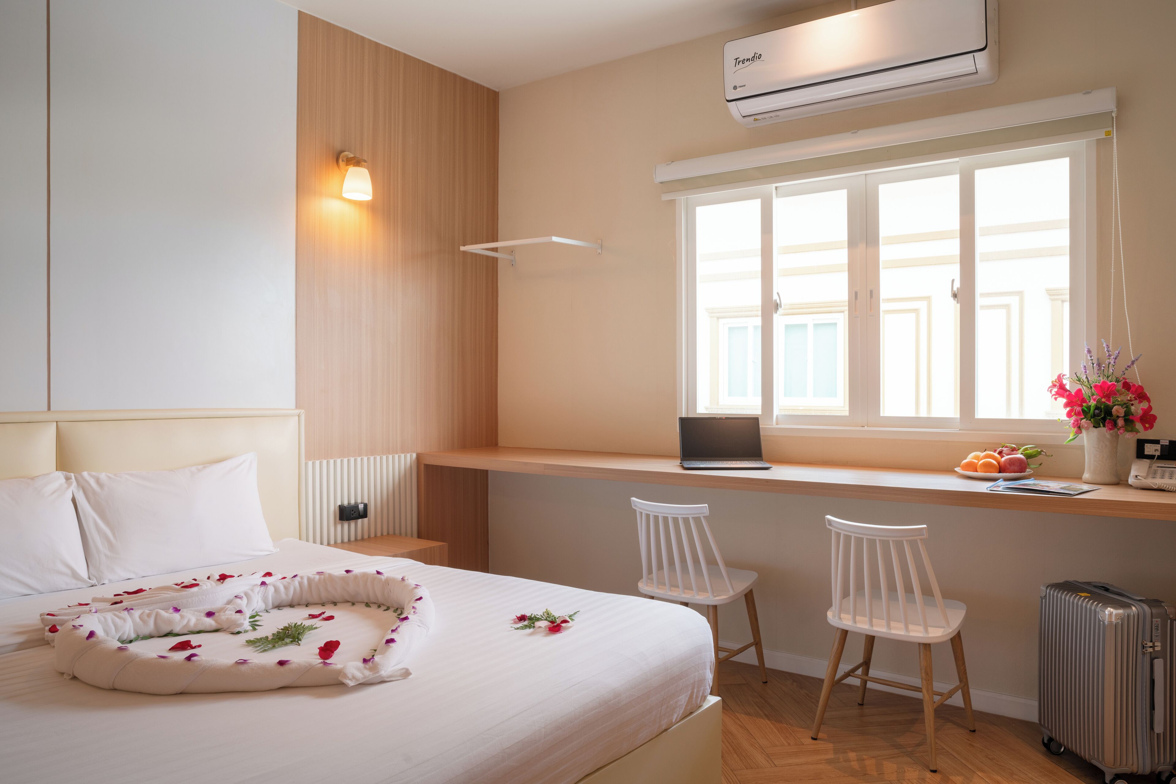 A hotel room with a bed, a desk with a laptop, two white chairs, a suitcase, and a window with a view.