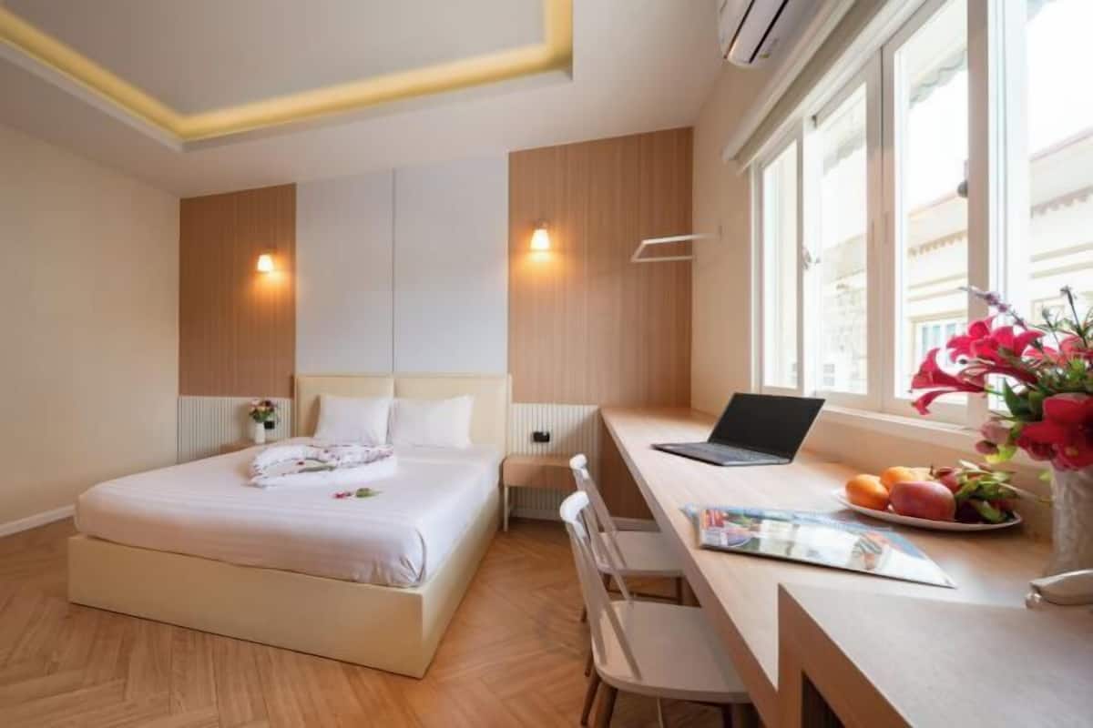 A hotel room with a bed, a desk with a laptop, a chair, a small table with fruit, and a vase with flowers.