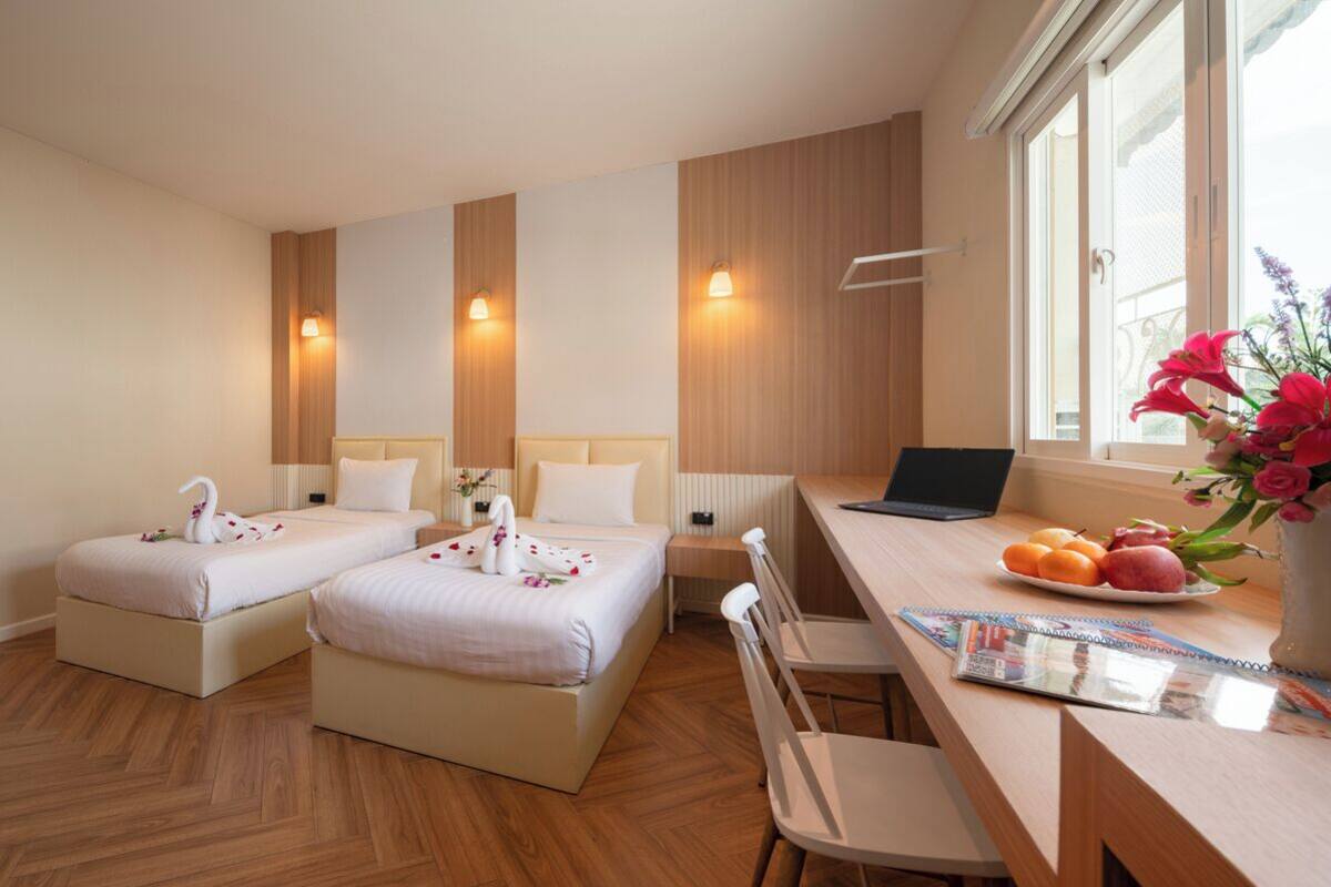A hotel room with two beds, a desk with a laptop, a vase with flowers, and a fruit bowl.