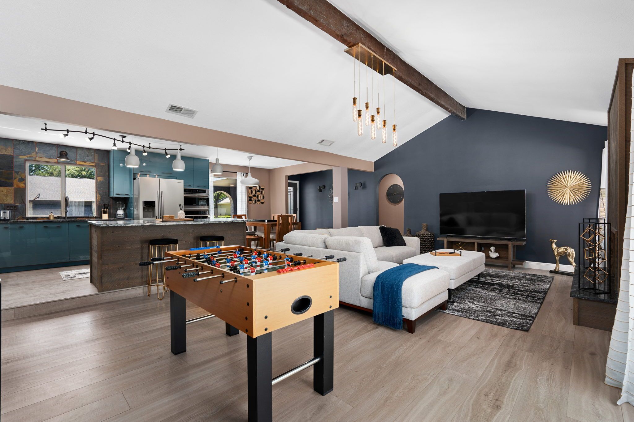 A modern living room with a foosball table, a sofa, a TV, and a kitchen in the background.