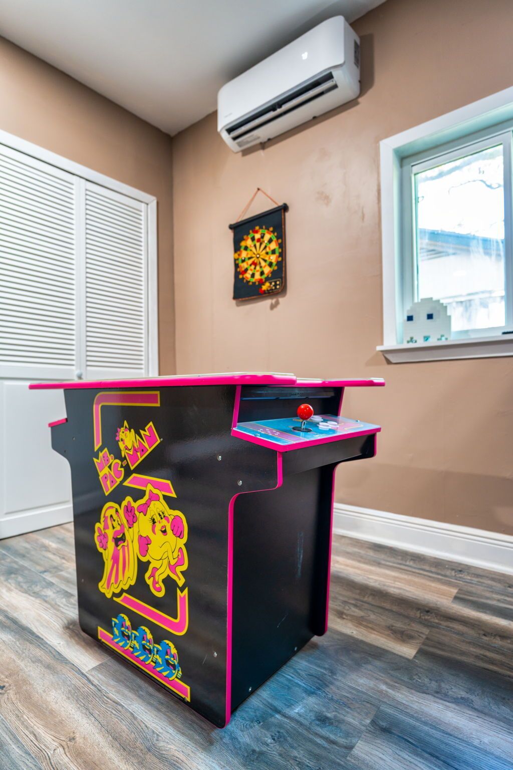 A room with a dartboard, an air conditioning unit, a window with a view, and a dartboard on the wall.