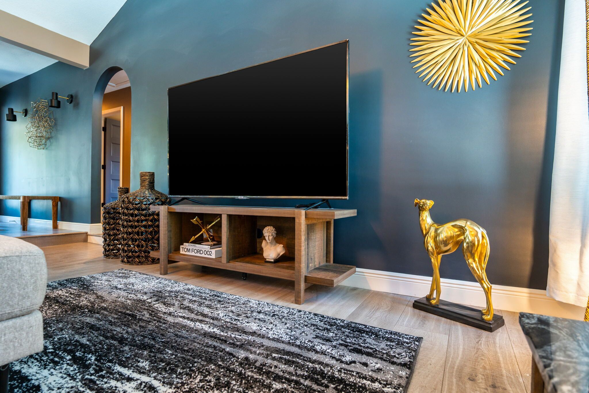 A modern living room with a large flat-screen TV on a wooden stand, a decorative golden statue, and a patterned rug.