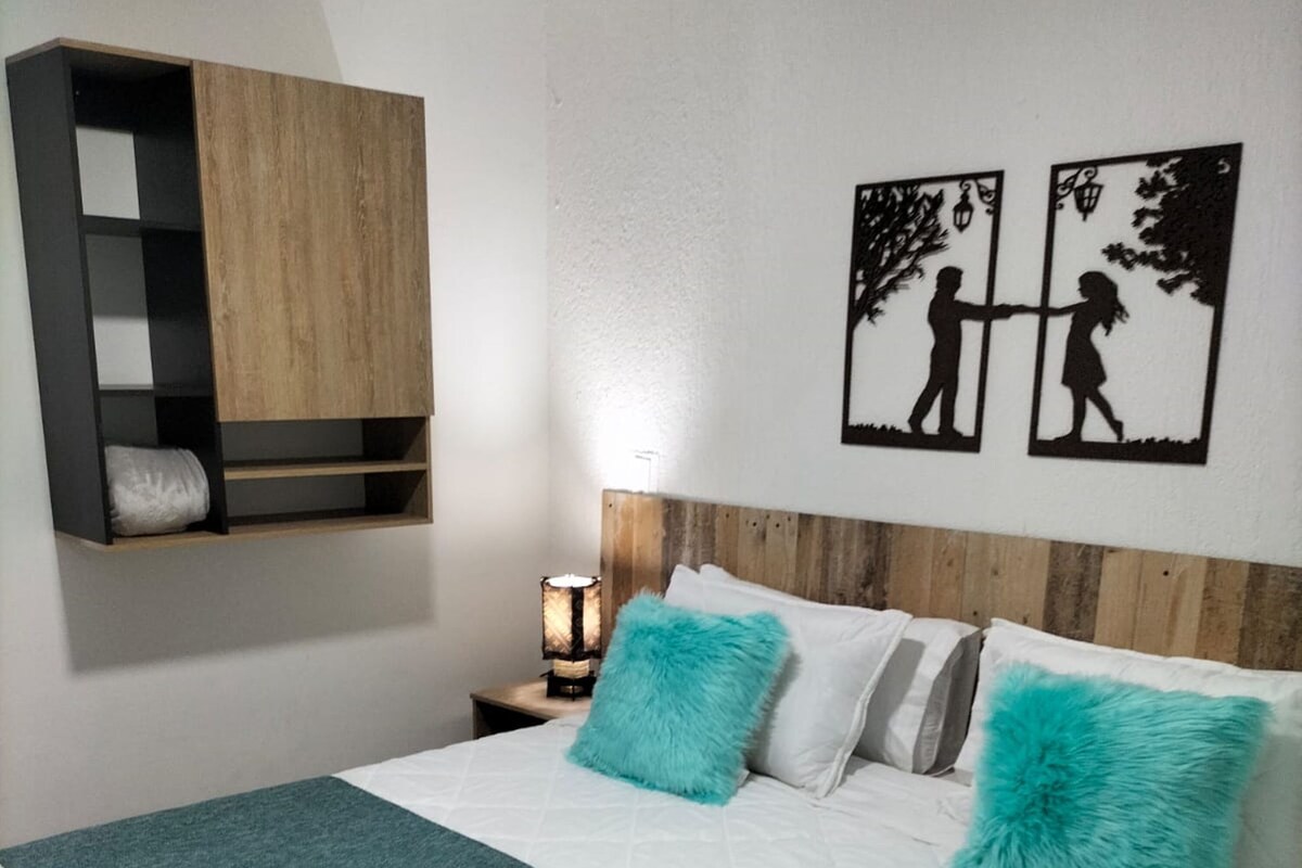 Ein ordentlich bezogenes Bett mit einer Türkis-farbenen Tagesdecke, einem Holzkopfschild und einem Nachttisch mit Lampe. Zwei Wandbilder zeigen spielende Kinder.